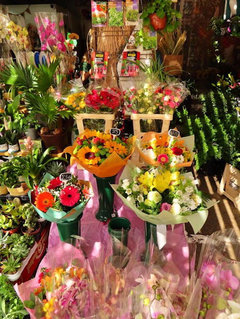A display of fresh floral bouquets and potted plants arranged on a pink table at a floral shop. The bouquets feature vibrant flowers such as sunflowers, gerbera daisies, and chrysanthemums in colors l