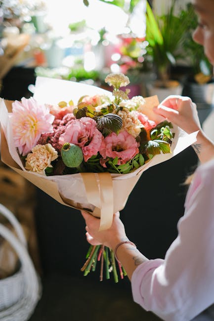 A person holding a large, freshly arranged floral bouquet wrapped in cream and white paper. The bouquet features pink peonies, cream carnations, purple statice, and green foliage, arranged in a lush, 