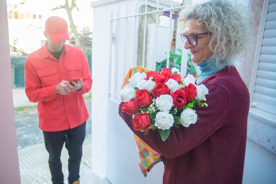 A woman with curly gray hair and glasses holding a vibrant bouquet of fresh red and white roses, wrapped in colorful paper, during a flower delivery at her doorstep. The arrangement is lush with green