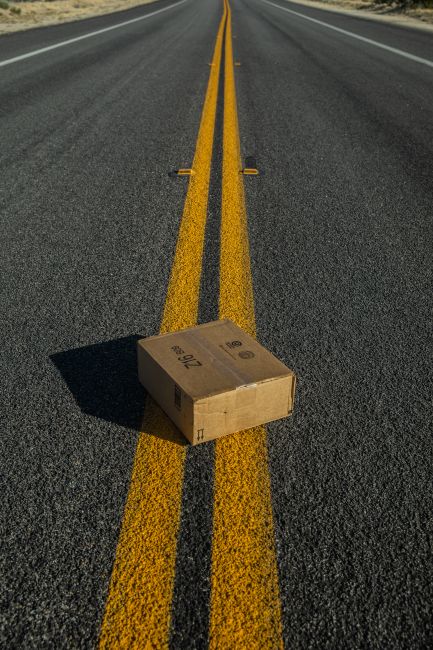 A cardboard delivery box is positioned in the middle of a straight, empty asphalt road with double yellow lines running down the center. The box has printed markings and appears sealed, casting a shad