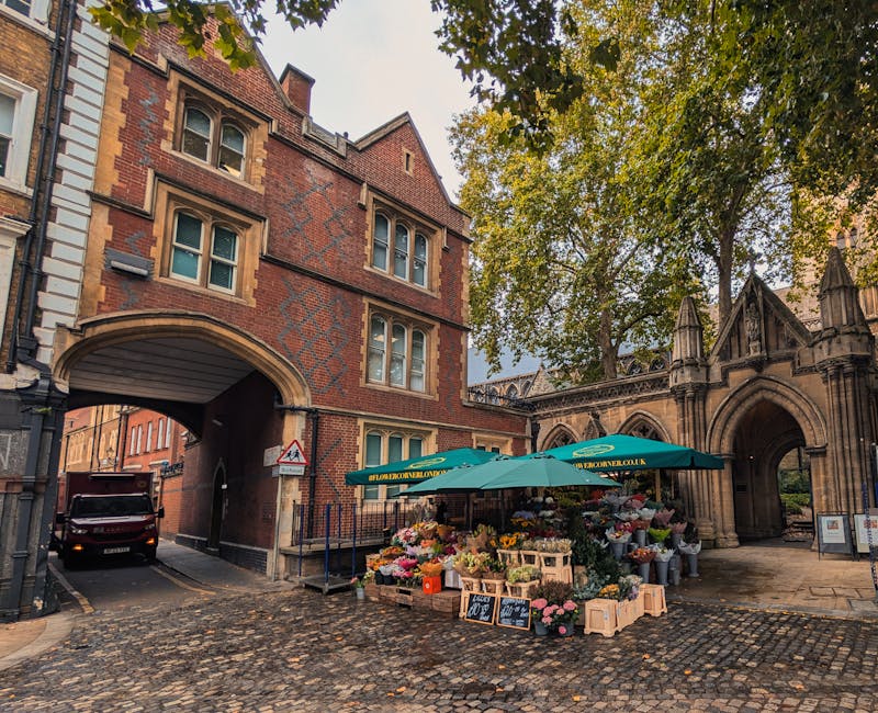 A vibrant flower stall set up outside a historic brick and stone archway in South Woodford, featuring a large green parasol with the [DOMAIN] logo. The stall displays a variety of fresh flowers includ