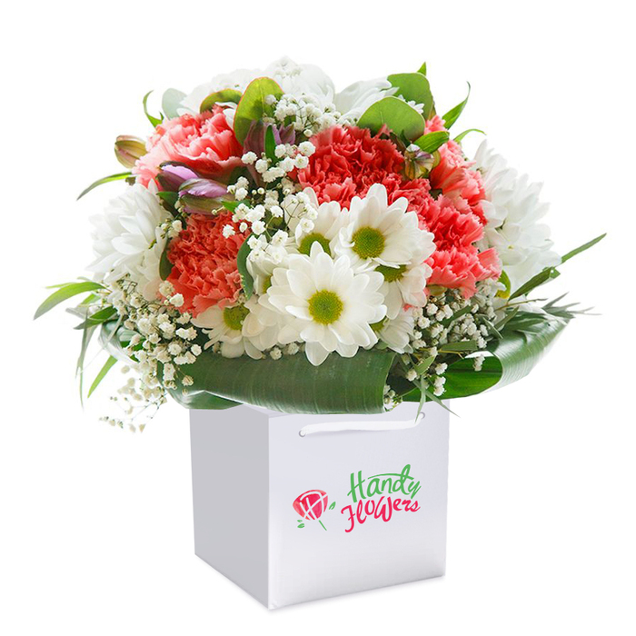 A vibrant floral bouquet, thoughtfully arranged by Flower Delivery South Woodford, rises from a sleek white square gift bag set atop a seamless white backdrop. Soft, deeply textured carnations in hues of coral, peach, and vibrant crimson unfurl their ruffled petals throughout the rounded ensemble, their delicate fragrance and lively colours conjuring the inviting ambiance of South Woodford's bustling George Lane. Scattered amongst them are pristine white daisies-each crowned with a luminous lime-green centre-contributing an element of cheerful radiance reminiscent of the area's welcoming parks and tree-lined streets. Wispy, cloud-like stems of baby's breath meander gently within the bouquet, weaving a light, airy tapestry throughout. Subtle pops of unopened, purple-tinged buds peek from behind rich green foliage, adding depth and intrigue, while a lush collar of broad, glossy leaves wraps protectively around the base, mirroring the thriving leaves seen in Churchfields Green. The bouquet's crisp white gift bag is finished with sturdy white ribbon handles, designed for easy, elegant transport-be it for a birthday surprise near South Woodford Station or marking an anniversary at home. This hand-crafted floral presentation radiates joy, refinement, and freshness, exemplifying true South Woodford floristry and making it an ideal gift for every heartfelt moment.