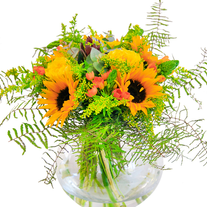 Stepping into the gentle light of a South Woodford morning, this vibrant bouquet radiates the very essence of late summer warmth. Sunflowers-resplendent in sun-kissed yellows and bold brown centres-stand tall beside lush yellow garden roses, whose tightly packed petals create swirling, velvety textures. Scarlet red Hypericum berries twinkle between the green leaves, like small jewels hidden among garden foliage. Fine sprays of fern fronds and airy, light-green filler branch out, adding feathery movement reminiscent of the soft wind in South Woodford's green spaces. Hints of eucalyptus introduce a touch of soothing coolness, balancing the arrangement with their dusty blue-green leaves. The bouquet is anchored in a crystal-clear, spherical glass vase, which elegantly displays the network of stems and the pure water they drink-offering a sense of clarity and freshness central to the florist's craft. The play of natural light enhances the bright colors, deep shadows, and each intricate detail. Deeply aromatic, the bouquet's scent is gently sweet and hints at open meadows nearby. Whether given to mark a significant occasion or simply to fill a living space with happiness, this arrangement is a joyful tribute to the everyday beauty found in South Woodford and the skilled hands that shape it.