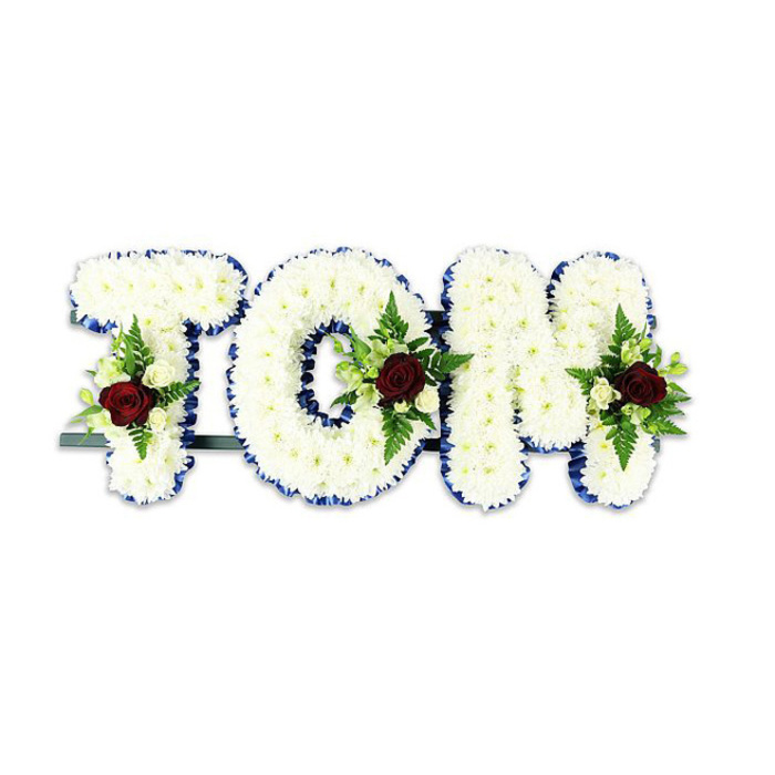 Handcrafted as a personal homage, the tribute spells TOM in voluminous three-dimensional letters formed from countless pristine white chrysanthemum blooms, each petal layered to build a soft, cushion-like surface. The letters sit against a stark white background that heightens the clarity of detail: the royal blue ribbon edging runs like tailored piping along each form, accentuating corners and curves with a bold, respectful frame. At the heart of each letter a composed spray balances the architecture-a singular, velvety deep red rose commands the gaze, surrounded by delicate ivory spray roses and tongued fern fronds whose green tones offer a cooling contrast and a faint, leafy scent. A subtle dark metal bar or support is visible beneath the arrangement, its practicality unobtrusive yet necessary to display the tribute upright at a service or beside a casket. The overall feeling is composed and steady, an expression of love and remembrance that reads clearly from the front pew: white for solace, blue for dignity, and red to mark the personal bond. As an experienced florist who often prepares memorials for families in and around South Woodford and nearby Wanstead Park, I choose materials and placement to ensure the letters feel both handcrafted and dignified, a calm, tactile statement that honours a life with understated beauty and care.