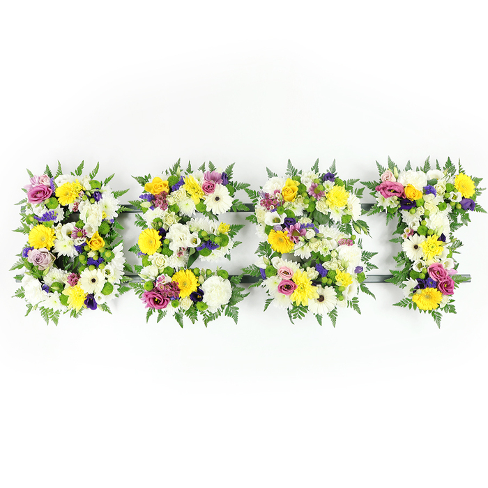 An elegant, statement floral tribute composed to spell BEST rests against a stark white studio background, each letter densely packed with freshly cut blooms and lush greenery. The dominant palette is uplifting: pure white chrysanthemum-like blooms form generous, soft mounds across the lettering, while bright yellow roses and gerbera daisies scatter sunlit notes through the design. Cooler violets come from clusters of purple lisianthus and lavender-hued small chrysanthemums that bring refinement and contrast, and vivid magenta spray roses or carnations are placed like affectionate punctuation throughout. Sprays of deep green fern fronds encircle and define the letters, their textured fronds creating a natural border that softens the geometry. A discreet dark green or metal frame is visible in places - notably across the horizontal bar of the E and near the T - indicating the careful internal support that allows such a dense composition to sit securely. The scene is evenly lit to showcase petal detail, freshness and the slight, sweet perfume of newly cut flowers. This refined tribute is well suited to funerals, memorial services or commemorative displays and could easily be chosen to honour a beloved family member or friend. As owners of Flower Delivery South Woodford, we craft these bespoke letter tributes for services across South Woodford, from deliveries to the chapel by the station to placements near the quieter corners of Wanstead Flats, always aiming for dignity, colour harmony and lasting freshness.