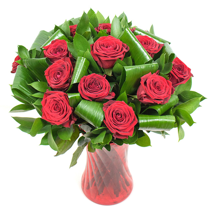 This image presents a deliberate, artful bouquet of more than a dozen deep crimson roses, each blossom at perfect bloom with tightly coiled, velvety petals that open to reveal a plush, fragrant centre. The colour is saturated and warm; under soft, even light the reds deepen to a tactile wine tone while petal edges gleam with delicate highlights. Abundant dark green foliage nests the blooms-slick, flat leaves providing a dense verdant field-and larger glossy leaves have been manipulated into elegant folds and spirals, like little green columns, lending structural support and contemporary flair to the classic roses. Those architectural leafy forms guide the eye upward and provide pleasing negative space between each crown of petals, balancing luxuriance with refined geometry. The bouquet is held within a tall, translucent ruby-red glass vase whose slender form and subtle ribbed twist create long vertical lines that echo the stems and catch reflections of light. The vase tapers at the waist and flares slightly at the lip, creating a graceful profile on a white studio background that isolates the composition and intensifies the contrast between the rich reds and deep greens. The overall effect is romantic and assured: a scent-forward, tactile arrangement that reads as both a gift for anniversaries and an elegant focal piece for a special dinner. Locally minded florists in South Woodford might place this on a mantel in a George Lane home or deliver it to a celebratory table near Roding Valley Park, where the bouquet's classic romance will feel perfectly at place.