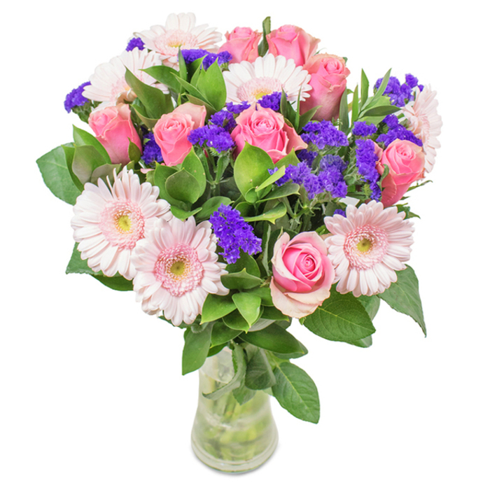 A vibrantly crafted bouquet presented in a clear glass vase, photographed against a crisp white backdrop-each element reflecting the attentive artistry found in South Woodford's independent flower shops. At the heart of the display, seven to nine pink roses-some tightly furled and others just beginning to open-showcase the classic spiral structure so beloved by rose enthusiasts. These are interlaced with five to seven pale pink gerbera daisies, their generous, almost white-centred faces radiating gentle warmth akin to the first blooms in Roding Valley Park after spring rain. Between these signature flowers, clusters of vivid purple statice add a striking pop of colour and textural contrast, their papery surfaces refracting light and lending an energy that enlivens the softer pastels. Rich, deep green foliage creates a lush frame at the base, heightening the sense of freshness and abundance while adding hints of a clean, green scent often experienced after a walk near South Woodford's greenspaces. The bouquet's carefully arranged stems are visible through the vase, resting in clear water that highlights their pristine condition-freshly cut and expertly selected for local flower delivery. Perfect for commemorating a birthday, anniversary, or Mother's Day, or simply as a delightful centrepiece for a home or office along Hermon Hill, the arrangement conveys a message of joy, celebration, and heartfelt affection.