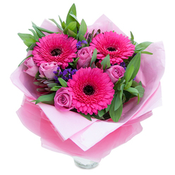 This composition presents three striking fuchsia-pink Gerbera daisies at its centre, each with dozens of crisp, radiating petals surrounding a dark, dramatic eye that anchors the arrangement visually. Interlaced around them are delicate roses in a softened lavender-pink or dusty-rose tone, their tight buds and unfolding curves suggesting freshness and a gentle perfume of rose. Sprigs of deep purple statice or limonium are tucked between the larger blooms, their tiny clustered florets providing airy texture and a cool counterpoint to the warm pinks. Lush green foliage-broad, glossy leaves that could be salal alongside the glaucous roundness of eucalyptus-creates a verdant backdrop, lifting the palette and framing each blossom with natural fullness. The bouquet is wrapped for gifting: an inner layer of pale pink, matte tissue paper supports the stems, while an outer veil of sheer, slightly crinkled fuchsia-pink cellophane adds a subtle shine and cohesive colour echo. The wrappings are folded with the meticulous hand of a neighbourhood florist, and the lower portion reveals the top lip of a clear glass vase, suggesting this is ready to be placed in a bright window or on a dining table. The overall mood is cheerful and affectionate, like a sunny visit to Roding Valley Park; it's the kind of arrangement Flower Delivery South Woodford would prepare for anniversaries, birthdays, or to send a heartfelt 'thank you'. Soft daylight in the photo enhances petal textures and the bouquet's inviting presence.