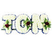 A personalised funeral flower arrangement spells out the name TOM in striking, three-dimensional letters, supported on a slim dark frame that disappears against the plain white background and allows the flowers to take centre stage. Each capital letter is densely based in immaculate white chrysanthemums, their rounded heads pressed closely together to create a soft, cushion-like surface that feels both gentle and strong. Around the outer edges, a neatly folded royal blue ribbon traces every corner and curve, its deep colour providing a dignified outline that brings to mind the calm, steady character of many farewell gatherings held around South Woodford's churches and chapels. At several points across the tribute, small sprays of richer colour draw the eye: clusters of deep red roses with velvety, layered petals, accompanied by small white or cream spray roses and slender white floral spikes similar to freesia. These blooms are set into pockets of lush green foliage and fine fern fronds that overflow slightly from the letters, introducing a natural, flowing movement to the otherwise structured design. The balance of white, blue, red, and green creates a harmonious, respectful tone, symbolising purity, remembrance, and enduring love. The whole piece feels like a carefully considered final gift from family or friends, the sort of bespoke name tribute a local family might arrange for same-day delivery through a South Woodford florist to honour Tom with grace, beauty, and a sense of personal connection during a time of loss.