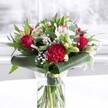 A luxurious red and white bouquet stands proudly in a clear cylindrical glass vase, the fresh green stems visible through the water like neat brushstrokes, suggesting careful preparation by an expert florist. Deep red roses with velvety, unfolding petals cluster at the heart of the design, their classic, romantic glow balanced by a large, ruffled crimson carnation at the front that adds rich texture and drama. Around them, soft white alstroemeria with speckled throats, creamy chrysanthemum-style blooms, and several slender white lily buds create a luminous contrast, as if pockets of light are tucked among the scarlet tones. Fine clouds of white gypsophila drift between the larger flowers, giving a sense of lightness and air, like gentle mist over Eagle Pond in South Woodford on a clear morning. At the base, broad, glossy dark green tropical leaves are wrapped and curved to form a polished collar, while pointed greenery and eucalyptus-like foliage thread upward, bringing layers of green that feel cool and refreshing to the eye. The bouquet rests on a spotless white surface, with a softly blurred, daylight-bright background that could easily be a sunlit window along George Lane. The lighting is even and kind, catching each petal and leaf so that the arrangement appears just-delivered, full of life and scent. It feels perfect for a South Woodford anniversary dinner, a thoughtful birthday surprise, or to welcome a new neighbour, offering warmth, admiration, and a refined sense of celebration in any home or office.