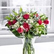 A vibrant bouquet of deep fuchsia-pink roses and ruffled carnations stands proudly in a clear glass vase on a crisp white tabletop, bathed in soft morning light from a nearby window. Each rose is lush and velvety, some fully open to reveal layered petals, others still swirling inward like a quiet promise, while the carnations add volume with frilled edges that catch the light. Nestled between the bold pinks, ivory alstroemeria unfold gently, their trumpet-shaped blooms edged with faint green, and a scattering of tiny white gypsophila, or baby's breath, brings a misty, cloudlike softness that lightens the arrangement. Large, glossy aspidistra leaves cradle the flowers, their broad, dark green surfaces curving around the base to create a graceful, structured silhouette that feels both contemporary and classic. Slimmer stems of lighter greenery weave upward, adding movement and depth so the arrangement appears almost to breathe. The glass vase, filled with clear water, reveals neatly cut stems lined up like a tidy chorus, reinforcing the sense of freshness and care. Behind the bouquet, the window view is gently blurred and muted, perhaps reminiscent of a quiet street in South Woodford early in the day, so that all focus remains on the glowing colours and textures of the flowers. As a florist familiar with homes along George Lane and near Eagle Pond, I can imagine this design lifting the mood of a breakfast table, brightening a birthday surprise, or serving as a heartfelt thank-you. The overall feeling is cheerful and optimistic, like a warm note of appreciation captured in petals and leaves, ready to be delivered to someone special in the local community.