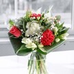 A contemporary red and white bouquet arranged in a clear glass vase, captured in soft, cool daylight as if placed by a bright South Woodford window overlooking George Lane. At the heart of the design, a coral-red rose opens its layered petals beside a ruffled crimson carnation, their rich tones pulsing with warmth and emotion. Around them, clusters of dainty white chrysanthemums and tiny daisy-like blooms add a cloud-soft lightness, their petals fine and feathery against the eye. Creamy white lilies, some just beginning to unfurl and others still in smooth, sculptural buds, rise gently through the arrangement, promising more beauty in the days ahead. Glossy, deep emerald leaves are folded and curved at the base, wrapping around the stems in graceful arcs that give the bouquet a modern, architectural silhouette. The glass vase reveals the straight green stems and clear water, hinting at the freshness of flowers that could have just arrived from a local market near Wanstead or Epping Forest. Light streaming through the window catches the sheen of each leaf and petal, creating subtle highlights and shadows that emphasise texture and depth. You can almost sense the fragrance: a clean, leafy freshness edged with soft sweetness from the lilies and rose. This floral composition feels uplifting yet calm, the sort of arrangement you might have delivered across South Woodford to celebrate a new home, mark a birthday, or simply bring a touch of everyday elegance to a kitchen table or office desk.
