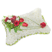 A solemn, meticulously crafted rectangular sympathy pillow composed entirely of hundreds of small, snow-white chrysanthemum blooms, the surface so densely packed it reads like a soft, clouded textile. Around the slightly raised edges a slim band of pale green ribbon and subtle foliage peeks out, giving the cushion a gentle framed finish and a whisper of fresh colour. Resting on the upper left corner is an intimate spray of deep red roses and burgundy carnations-roses in both tight bud and full bloom whose velvety petals offer a warm, timeless contrast to the pristine white base. Tiny clusters of delicate white accent blooms-freesia or small spray chrysanthemums-nestle between the reds, catching the light and adding a quiet softness. Lush green leaves and feathery fern fronds create a verdant backdrop, while long, bright green bear-grass strands or decorative stems arc gracefully from the main cluster across the chrysanthemum field toward the lower right. Those sweeping green lines culminate in a single perfect red rose placed gently on the right side, as if carried there by memory; the composition reads as a visual line of thought, moving from collective comfort to the intimate sign of love. The arrangement is photographed against soft, neutral light that highlights texture and depth without drama, producing a calm, reverent mood. As a local florist familiar with South Woodford funerals and services near George Lane and Epping Forest, I would recommend this pillow as a dignified expression of condolence-quiet, tactile, and enduring-suitable for wakes, memorials or a graveside tribute where comfort and respect are paramount.