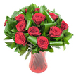 A luxurious, rounded bouquet of a dozen-plus deep crimson roses fills the frame, each bloom opened to reveal tightly spiraled, velvety petals whose colour reads almost wine-dark in the soft, even studio light. The roses sit closely clustered, their rich red heads forming a plush, tactile mass that suggests warmth and intimate celebration. Around and between the blooms, generous swathes of dark green foliage-glossy, flat leaves like Ruscus or Salal-create a dense, verdant backdrop that makes the reds sing. Larger emerald leaves, reminiscent of Aspidistra, have been expertly curled and rolled into sculptural loops and spirals; those artful green arcs provide architectural contrast and a modern edge while cradling each rose head delicately. The whole composition is anchored in a tall, translucent ruby-red glass vase, its surface subtly ribbed or twisted so vertical lines catch the light and echo the stems inside. The vase narrows slightly at the waist before flaring gently at the neck, lending the arrangement an elegant silhouette suitable for a mantelpiece or dining centre. Set against a clean white background, every hue and texture reads clearly-the velvet of petals, the glossy sheen of leaves, the glass's polished glow-inviting the imagination to smell the classic, soft rose fragrance. As a local florist would suggest for anniversaries or Valentine's Day, this bouquet balances timeless romance with contemporary structural detail, a piece that speaks quietly of passion, thoughtful design, and a celebratory presence fit for a special table or a heartfelt gift delivered across South Woodford.