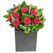 In this photograph a compact, modern bouquet of deep crimson roses takes centre stage, each bloom unfurling in generous, velvety spirals that catch the light like rubies against a satin ground. The roses are gathered tightly into a low, square matte black cube, its crisp geometry giving the composition a grounded, architectural presence. Around and between the heads of bloom, broad glossy green leaves have been folded and looped with deliberate care, their smooth surfaces and curled tips creating a rhythmic sense of movement and layered depth that frames every petal. The studio lighting is clear and steady, skimming the high points of petals and leaves so the roses glow with soft highlights while the foliage reads as bright, almost jewel-like emerald. Up close you can imagine the tactile contrast-the silk of the petals against the firmer, almost waxy texture of the greenery-and the subtle scent: classic rose warmed into a fresher, leaf-scented top note. As a local South Woodford florist I see this arrangement working as a quiet but decisive statement for anniversaries, a romantic surprise, or as an elegant focal piece for a small dining table by George Lane; it feels at once intimate and urbane. The design's restrained palette and confident structure make it suitable for both heartfelt declarations and refined interior styling, a bouquet that offers a tactile, fragrant reminder of careful craft and considered taste in every folded leaf and open bloom.